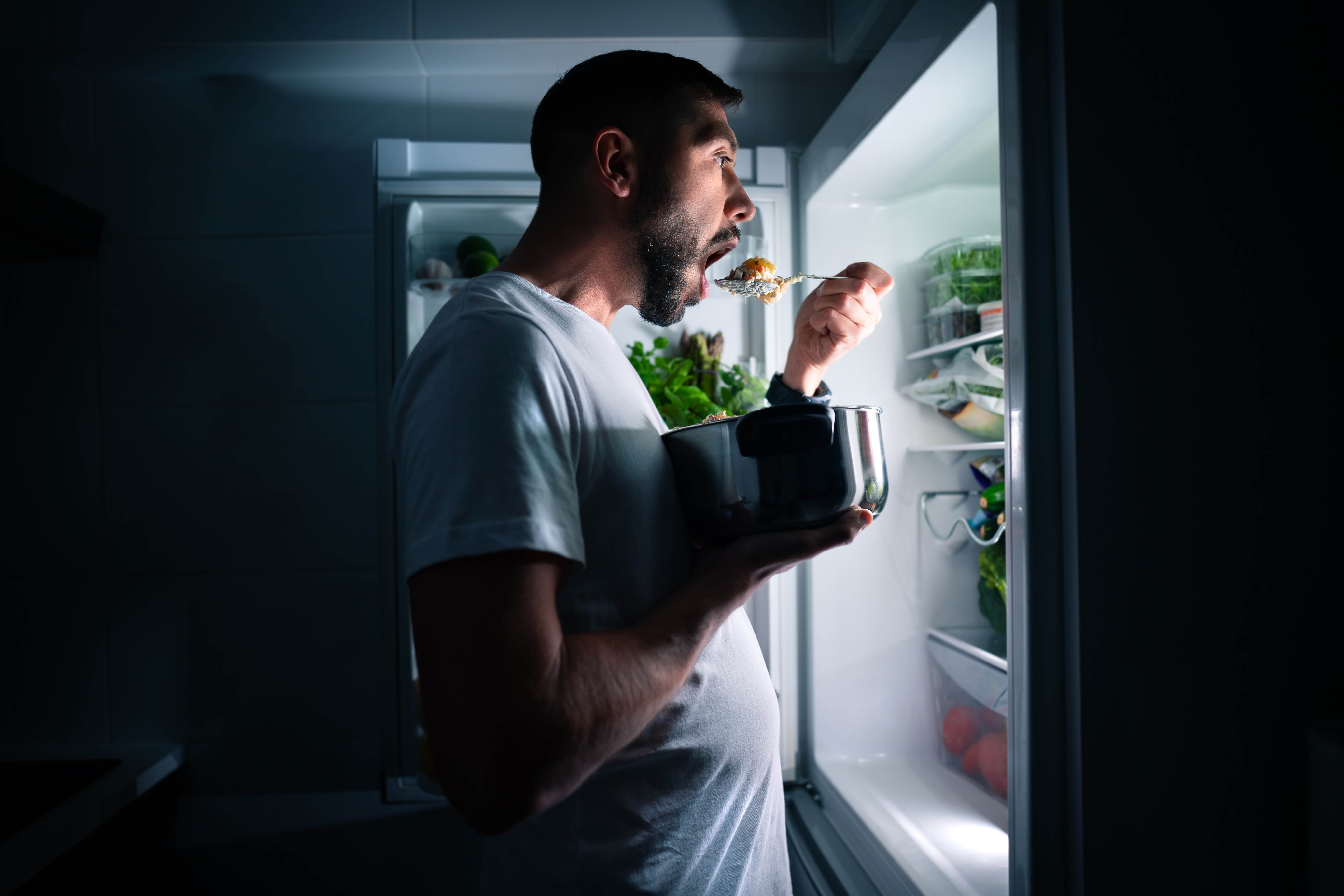 Man standing and eating with the fridge door open and the fridge light on him